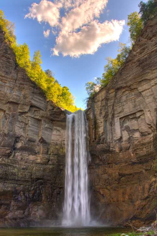 A stunning waterfall cascades down rocky cliffs, surrounded by lush greenery and a bright blue sky with fluffy clouds.