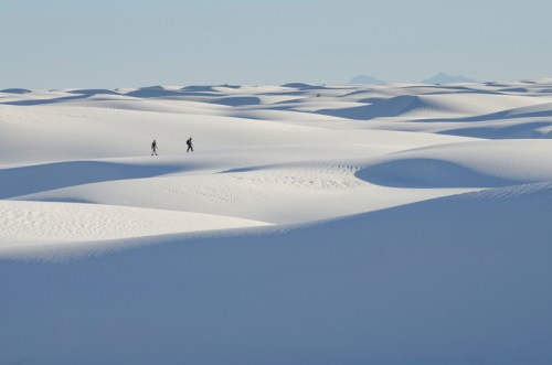 Two hikers traverse a vast, undulating landscape of white sand dunes under a clear blue sky.