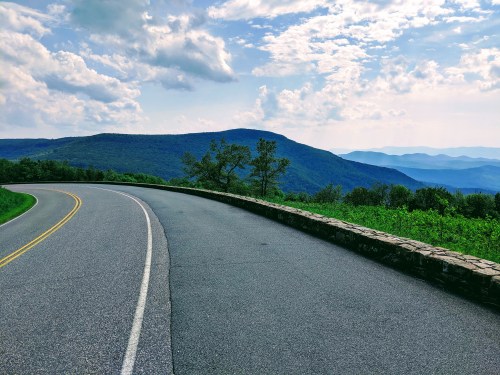 A winding road curves through lush green hills under a partly cloudy sky, with distant mountains in the background.