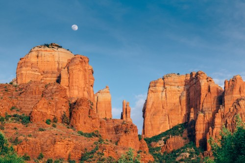 A scenic view of red rock formations under a blue sky with a crescent moon in the background.
