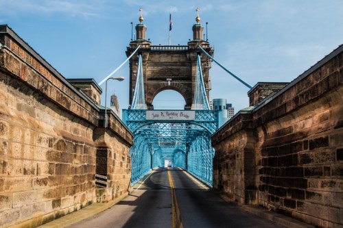 A view of the John A. Roebling Bridge, featuring its blue suspension cables and stone supports, leading into a cityscape.
