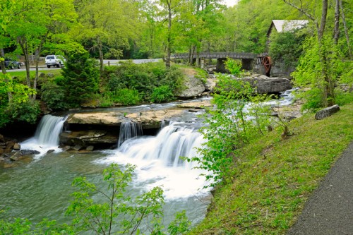 A serene scene of a flowing river with small waterfalls, surrounded by lush greenery and a rustic building nearby.