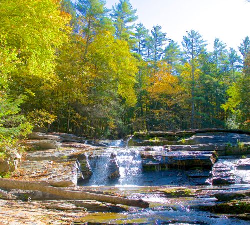 A serene river flows over rocks, surrounded by vibrant autumn trees under a clear blue sky.