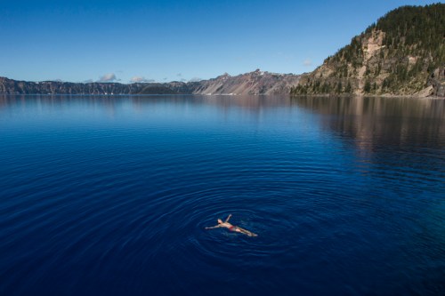 A person swims in the calm blue waters of a lake surrounded by mountains and clear skies.