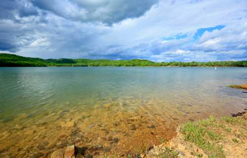 A serene lake surrounded by green hills under a cloudy sky, with clear water revealing a rocky shoreline.