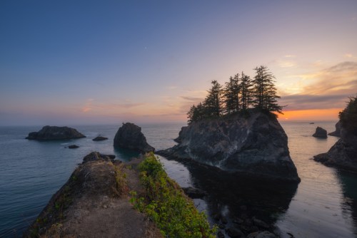 A serene coastal scene at sunset, featuring rocky outcrops and a small island with trees, reflecting in calm waters.