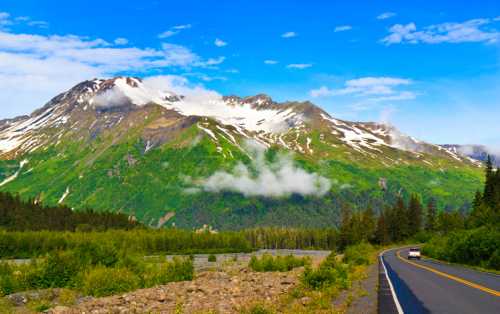 A scenic view of snow-capped mountains under a blue sky, with a winding road and lush greenery in the foreground.