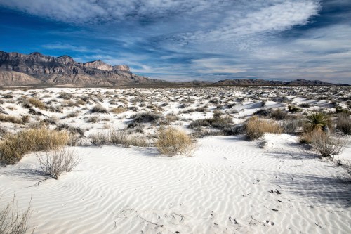 A vast desert landscape with white sand dunes, sparse vegetation, and distant mountains under a cloudy sky.