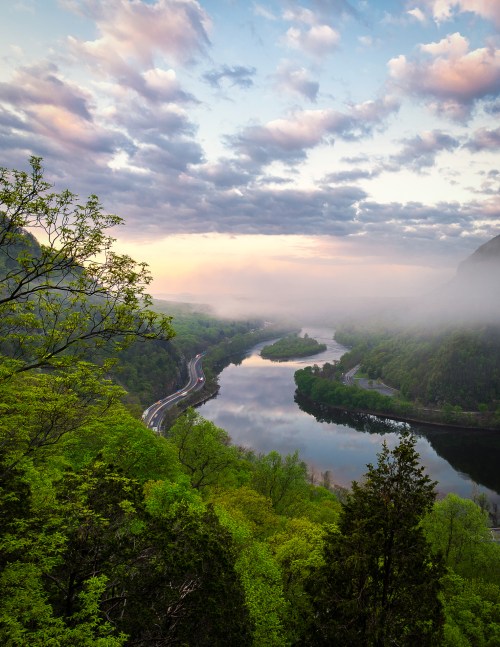 A serene river winds through lush green hills under a cloudy sky, with mist rising from the water at dawn.
