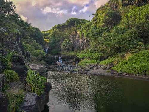 A serene landscape featuring a calm pool surrounded by lush greenery and a waterfall in the background under a cloudy sky.
