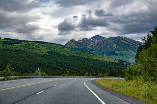 A winding road leads through lush green hills and mountains under a cloudy sky.