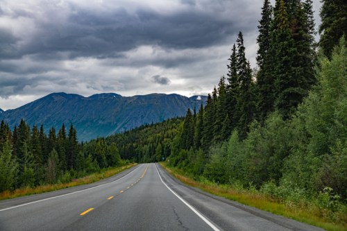 A scenic highway lined with trees, leading towards mountains under a cloudy sky.