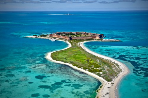 Aerial view of a tropical island with sandy shores, lush greenery, and clear blue waters surrounding a historic building.