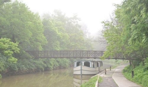 A foggy scene of a boat docked by a tree-lined canal, with a wooden bridge in the background.