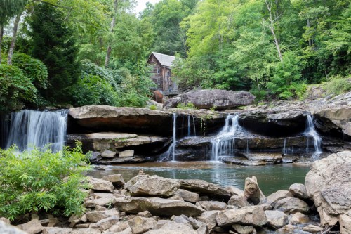 A serene landscape featuring a rustic mill by a waterfall, surrounded by lush greenery and rocky terrain.