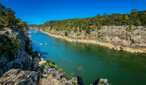 A serene river flows between rocky banks, surrounded by lush greenery under a clear blue sky.