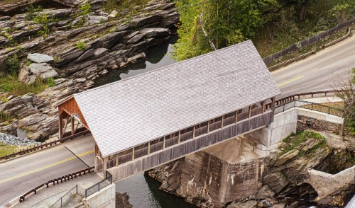A wooden covered bridge spans a river, surrounded by rocky terrain and greenery, with a road running alongside it.