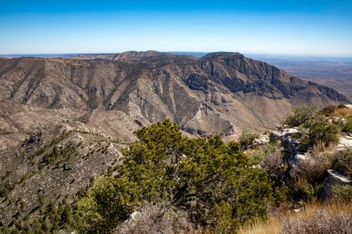 A panoramic view of rugged mountains and valleys under a clear blue sky, with sparse vegetation in the foreground.