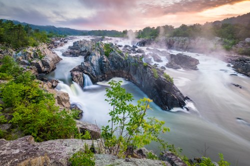 A scenic view of a river with cascading waterfalls, surrounded by lush greenery and misty cliffs at sunrise.