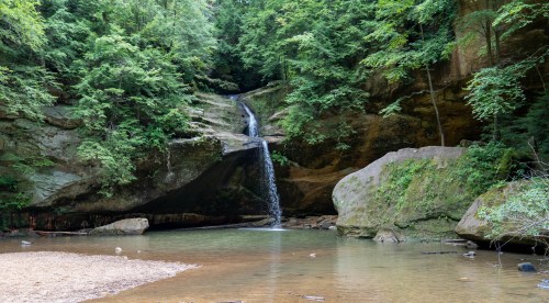 A serene waterfall cascades into a calm pool, surrounded by lush green trees and rocky cliffs.