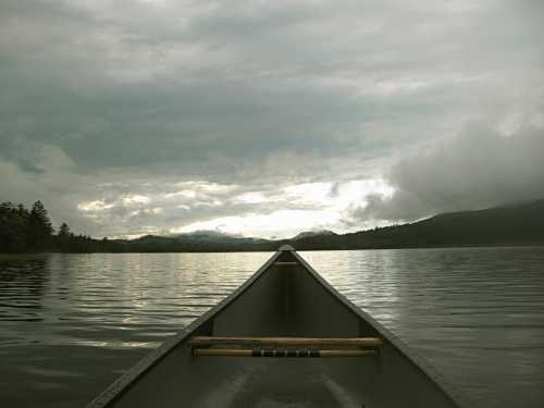 A canoe's bow on a calm lake under a cloudy sky, surrounded by distant mountains and trees.