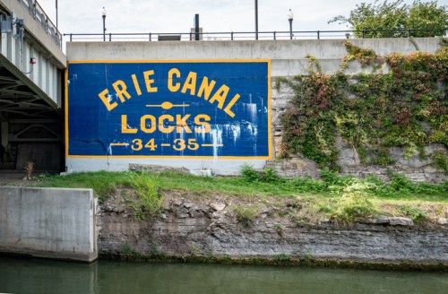 Sign on a wall reading "Erie Canal Locks 34-35," with greenery and water in the foreground.