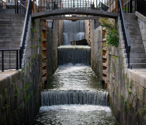 A serene waterway with cascading waterfalls, flanked by stone walls and a bridge overhead.