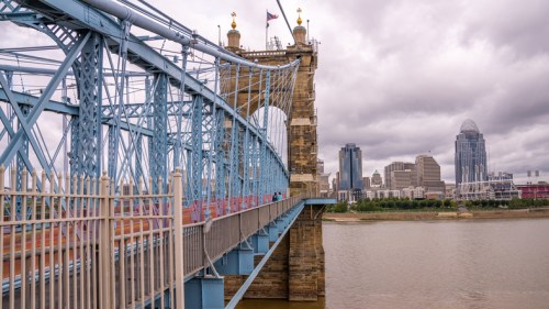 A blue suspension bridge spans a river, with a city skyline and cloudy sky in the background.