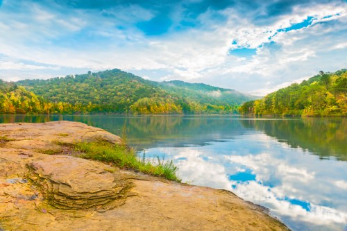 A serene lake surrounded by colorful autumn trees and mountains, reflecting clouds in the calm water.