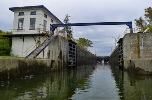 A view of a water lock with a control building, surrounded by calm water and greenery under a cloudy sky.