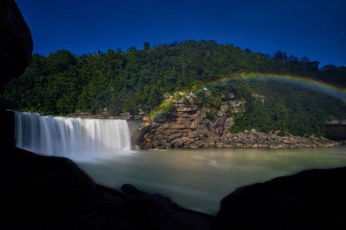 A serene waterfall cascades into a calm river, with a vibrant rainbow arching over lush green hills under a starry night sky.