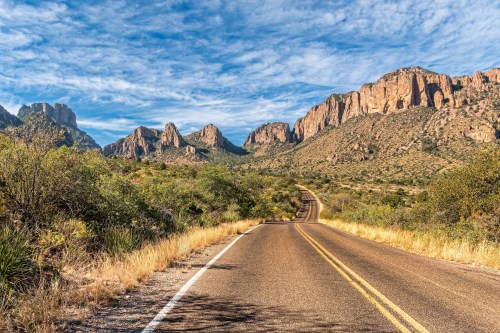 A winding road leads through a desert landscape with rugged mountains and blue skies in the background.
