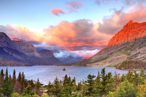 A serene lake surrounded by mountains, with vibrant sunset colors reflecting in the water and lush trees in the foreground.