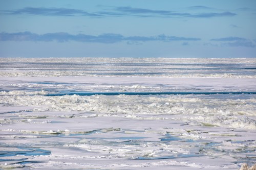 A vast icy landscape with fragmented ice floes under a clear blue sky and scattered clouds.
