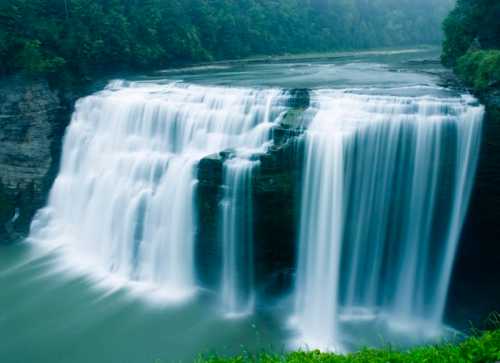 A serene waterfall cascading over rocky cliffs into a calm river, surrounded by lush greenery and misty atmosphere.