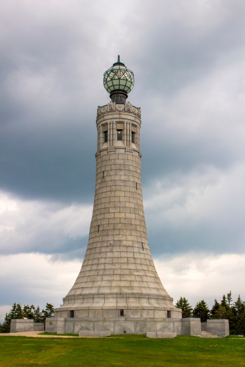 A tall stone tower with a glass lantern on top, set against a cloudy sky and green grass.