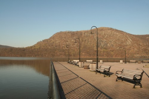 A peaceful riverside park with benches and lampposts, overlooking a calm waterway and a distant wooded hill.