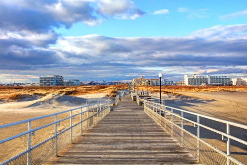 A wooden boardwalk leads to a beach, with dunes and buildings under a cloudy sky.