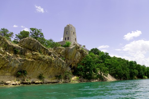 A stone tower atop a rocky cliff surrounded by lush greenery and a clear blue lake under a bright sky.