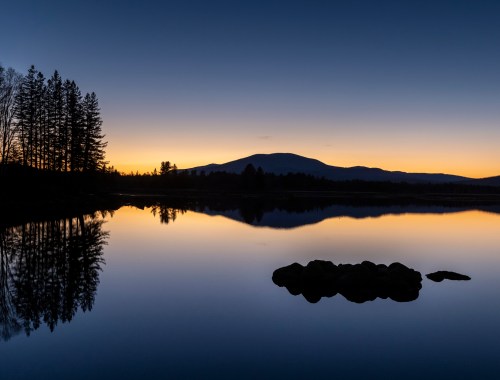 A serene lake at dusk, reflecting trees and a mountain silhouette against a gradient sky.