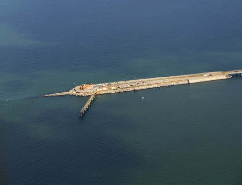 Aerial view of a long pier extending into calm blue waters, with a small building at the end and rocky edges.