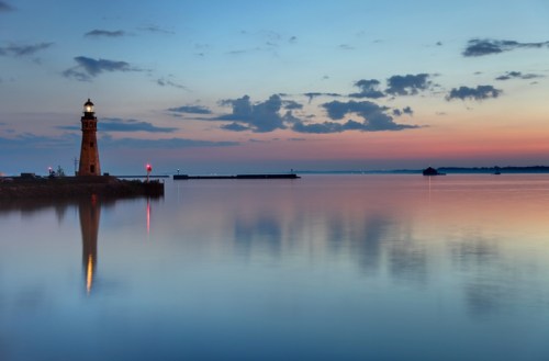 A serene sunset over calm waters, featuring a lighthouse and soft clouds reflecting in the water.