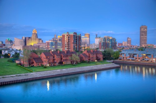 A scenic view of a city skyline at dusk, reflecting on a calm river with green spaces and modern buildings.