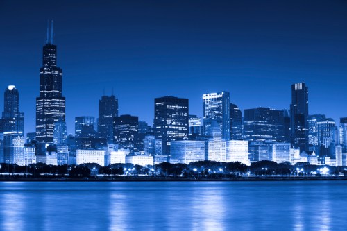 A nighttime city skyline with blue tones, featuring tall buildings and reflections on the water.