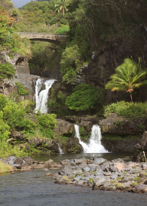 A scenic view of waterfalls cascading into a rocky stream, surrounded by lush greenery and a stone bridge above.