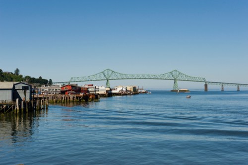 A calm waterfront scene featuring a green bridge, boats, and buildings along the shore under a clear blue sky.