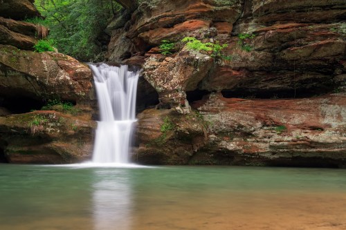 A serene waterfall cascades over rocky cliffs into a calm, clear pool surrounded by lush greenery.