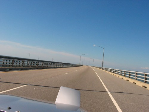 A view of a long, straight road on a bridge under a clear blue sky, with water visible on both sides.