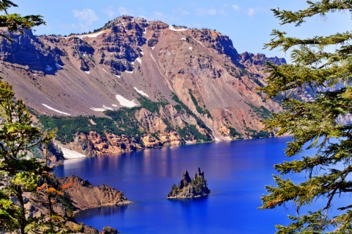 A scenic view of a blue lake surrounded by mountains, with a small island in the water and trees in the foreground.