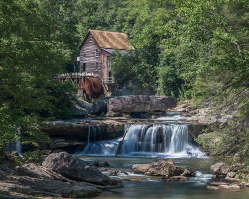 A rustic wooden mill beside a waterfall, surrounded by lush greenery and rocky terrain.